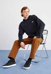 Svart hoodie med en framficka och broderad logotyp, tillsammans med bruna kordbyxor och svarta sneakers med vita sulor, sittande på en stol.