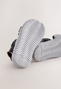 Sandales noires avec une semelle texturée, dotées de sangles réglables et d'un accent marron. La semelle est blanche avec un motif ondulé et des taches.