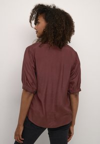 Burgunderfarbene Bluse mit Puffärmeln, glatter Textur und geknöpften Manschetten. Abgerundeter Saum und dezenter Glanz, seitlich betrachtet.