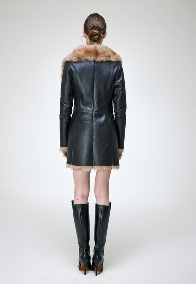 Femme aux cheveux en chignon portant un manteau en cuir noir avec bordure en fourrure et bottes noires à talons montant jusqu'aux genoux, debout, dos tourné sur un fond uni.