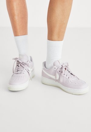 Ben iført hvide ribbede crew-sokker og lyserøde Nike Air Force 1 sneakers med hvide såler og snørebånd på en hvid gulv.