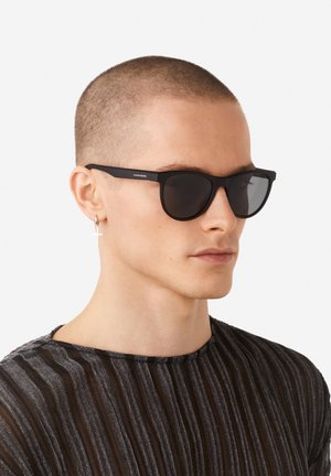 Melni saulesbrilles ar matētu apdari, apaļiem ietvariem un tumšiem stikliņiem. Uz kājiņas redzams zīmola nosaukums "Hawkers". Modelis valkā svītrainu kreklu.