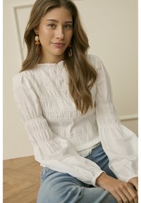 Junge Frau mit langen braunen Haaren, die eine weiße, geraffte Bluse und blaue Jeans trägt, sitzt und lächelt leicht vor einem neutralen Hintergrund.