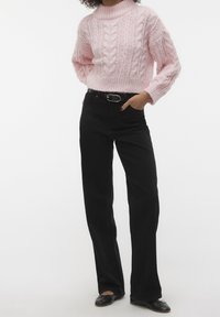 Personne portant un pull rose à torsades, un pantalon noir taille haute avec ceinture, et des ballerines noires, debout les mains dans les poches.