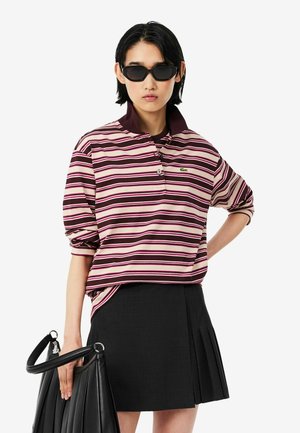 Personne portant des lunettes de soleil noires, une chemise à manches longues rayée rose et bordeaux, une jupe plissée noire et tenant un sac à main noir.
