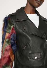 Giacca di pelle nera con chiusura a zip e diverse tasche. La manica sinistra presenta pelliccia sintetica colorata in tonalità di rosso, verde e blu.