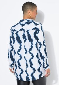 Koszula z długim rękawem z białym tłem i niebieskim falistym wzorem tie-dye, wykonana z lekkiego materiału, z klasycznym kołnierzykiem i zapinanymi mankietami.