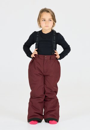 Pantaloni da neve bordeaux con ampi plissettamenti, bretelle nere regolabili con logo. Indossati sopra una maglietta nera a maniche lunghe e calze rosa.