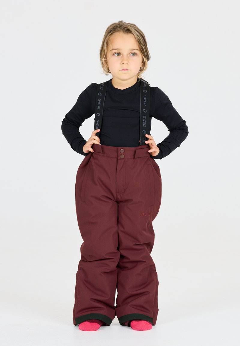 Pantaloni da neve bordeaux con ampi plissettamenti, bretelle nere regolabili con logo. Indossati sopra una maglietta nera a maniche lunghe e calze rosa.