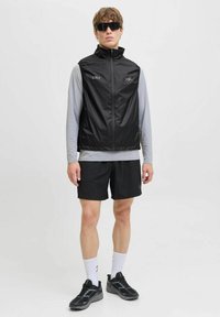 Zwarte lichte vest met hoge kraag, logodetail, gecombineerd met een grijs longsleeve shirt, zwarte shorts en sportief schoeisel.