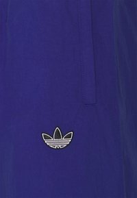 Tessuto viola con una trama liscia, caratterizzato da un logo Adidas ricamato in bianco e nero. Il design è semplice con dettagli minimi.