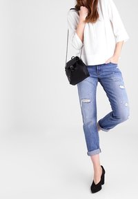 Biały, luźny top z drapowanym dekoltem; jasnoniebieskie, przetarte dżinsy; czarna torba typu bucket; czarne szpilki; swobodna poza.