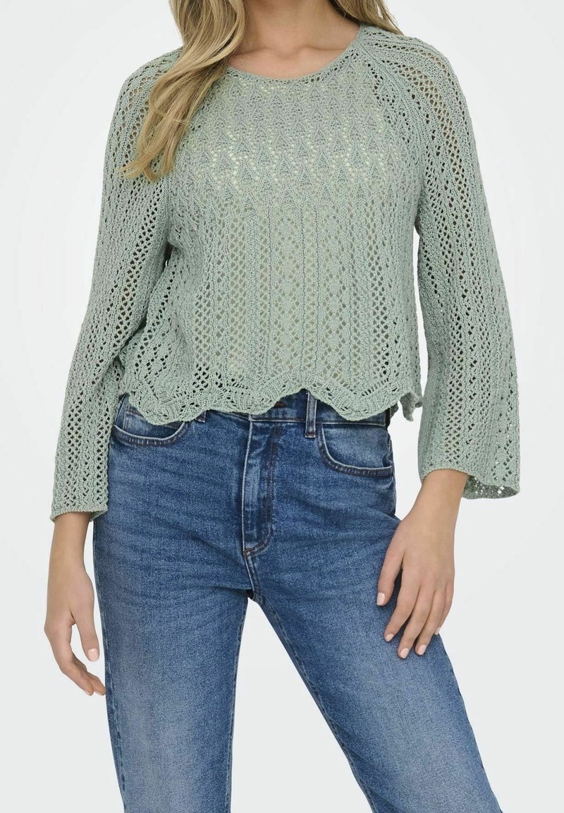 Pull vert clair en tricot avec un motif texturé, manches évasées et un ourlet festonné, associé à un jean bleu taille haute.