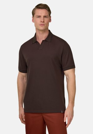 Homme portant un polo à manches courtes marron foncé et un pantalon couleur rouille, debout devant un fond blanc uni.