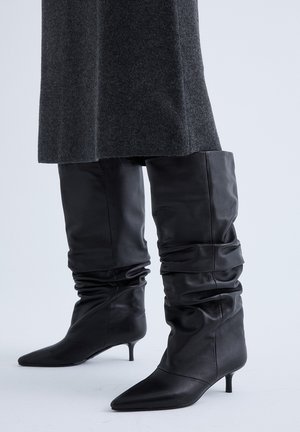 Botas negras de cuero holgadas hasta la rodilla con punteras puntiagudas y tacones bajos tipo kitten, usadas con una falda o vestido gris oscuro.