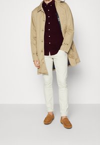 Gabardina beige sobre una camisa de botones de pana burdeos, combinada con pantalones color crema y mocasines de ante marrón.