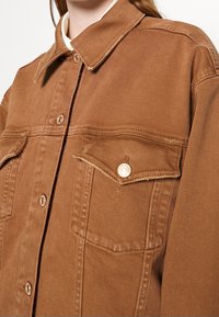 Veste en denim marron avec un devant à boutons, deux poches poitrine et un col classique. Elle présente des boutons en métal et un tissu texturé.