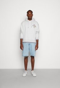 Felpa grigio chiaro con cappuccio e logo ricamato, abbinata a pantaloni corti in denim blu con orli sfilacciati e scarpe da ginnastica bianche con dettagli grigi.