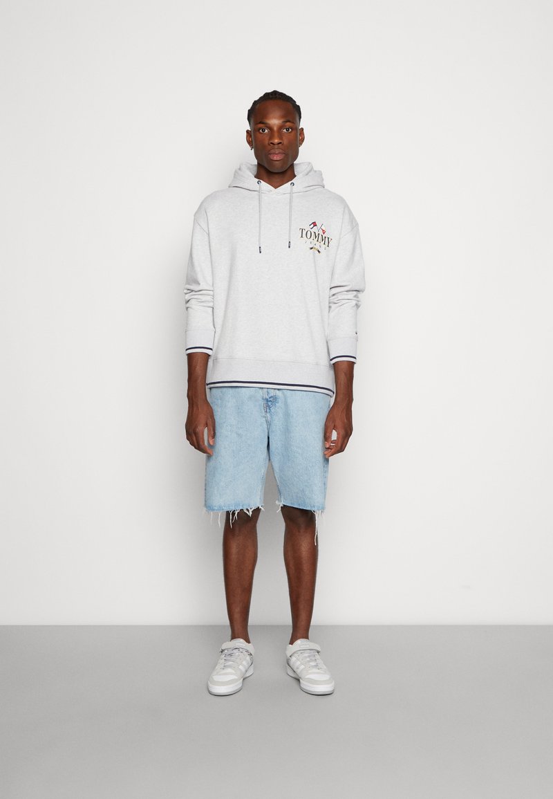 Felpa grigio chiaro con cappuccio e logo ricamato, abbinata a pantaloni corti in denim blu con orli sfilacciati e scarpe da ginnastica bianche con dettagli grigi.