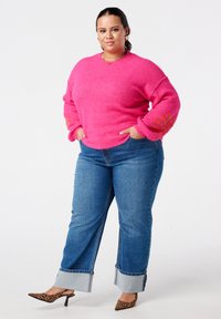 Vrouw staand met handen in de zakken, gekleed in een felroze trui met oranje bloemenborduursel, opgerolde blauwe spijkerbroek en schoenen met luipaardprint.