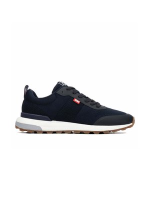 Zapatilla deportiva negra con suela blanca, banda de rodadura marrón, cordones negros y etiqueta roja en el lateral, vista desde el lado exterior sobre un fondo blanco.