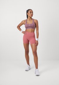 Športni nedrček v mauve barvi in visoko pasne hlače v svetlo roza. Model nosi bele športne čevlje in zapestnice. Gladek, raztegljiv material.