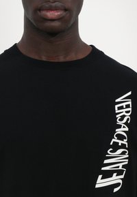 Svart bomull T-skjorte med rund hals, med fet hvit "VERSACE" tekst trykket vertikalt på venstre side. Glatt tekstur.