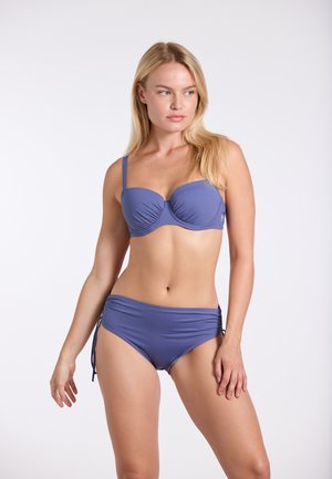 Woman with long blonde hair wearing a purple underwire bikini top and matching high-waisted bottoms, standing against a plain white background.