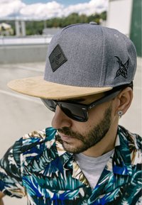Siva snapback kapa s bež vizorom, s crnim emblemu. Nosena s tamnim sunčanim naočalama i košuljom s višebojnim tropskim uzorkom.