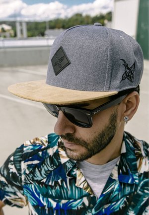 Szürke snapback sapka, bézs karimával, fekete emblémával. Sötét napszemüveggel és több színben pompázó trópusi mintás inggel viselve.