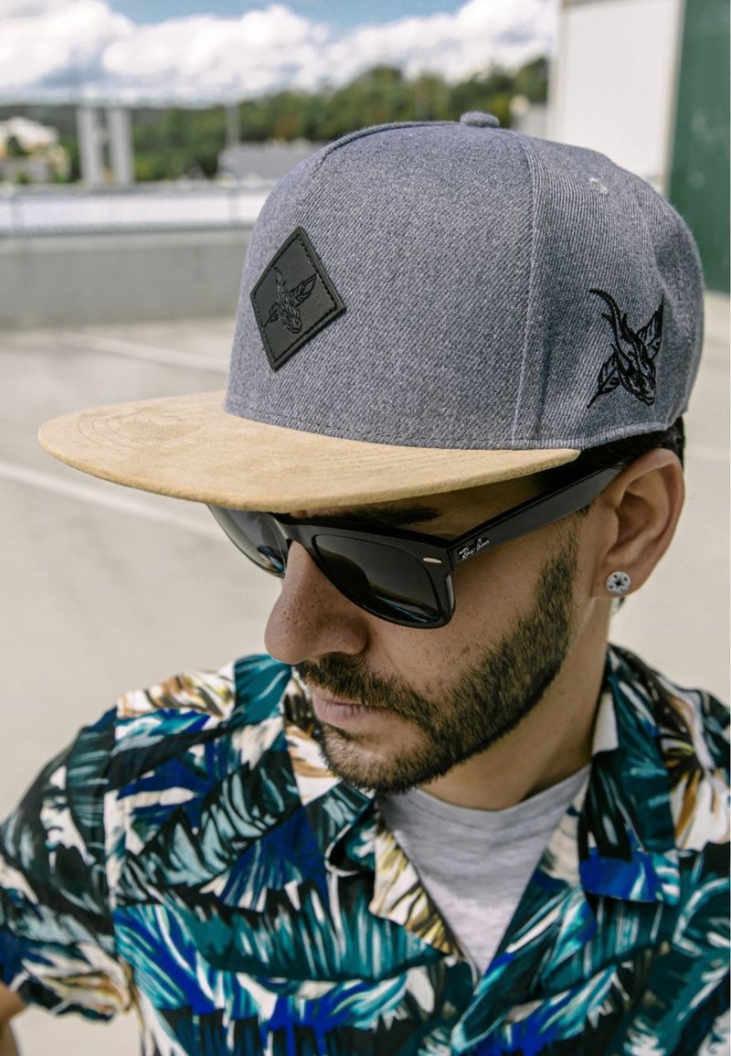 Siva snapback kapa s bež vizorom, s crnim emblemu. Nosena s tamnim sunčanim naočalama i košuljom s višebojnim tropskim uzorkom.