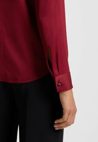 Blusa in satin rosso con maniche lunghe, caratterizzata da uno spacco e un dettaglio con bottone al polsino. Texture liscia e design elegante.