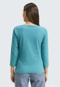 Türkises Langarmshirt mit weitem Rundhalsausschnitt und figurbetonter Silhouette. Sanfter, glatter Stoff. Mit geradem Saum und Dreiviertelärmel.