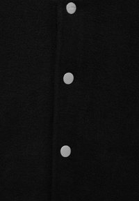 Gros plan sur un tissu noir texturé avec trois boutons-pression ronds argentés espacés uniformément, le bouton supérieur gravé du nom de la marque.