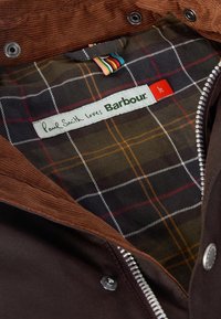 A barna kordbársony dzseki bélésének kockás mintázata sötétzöld, piros és fehér színű. Tartalmazza a "Paul Smith loves Barbour" címkét és kontrasztos cipzárt.