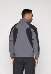 Αεροδυναμικό μπουφάν σε γκρι με μαύρες λεπτομέρειες, που διαθέτει ψηλό γιακά, ελαστικούς μανσέτες και απαλό ύφασμα. Η πλάτη έχει σχεδιασμό με πάνελ.