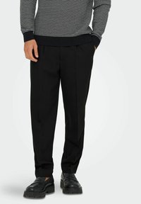 Schwarze maßgeschneiderte Hose mit glatter Textur, mit vorderen Falten und entspanntem Schnitt, kombiniert mit schwarzen Slippeschuhen.