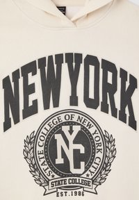 Felpa beige con testo nero audace "NEW YORK" e un grafico circolare con "STATE COLLEGE OF NEW YORK CITY" e accenti di alloro.