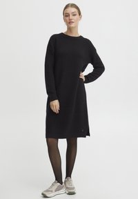 Abito in maglia nera con un design textured, maniche lunghe e spacco laterale. Abbinato a sneakers beige e collant neri.