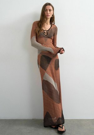 Vrouw draagt een lange, gebreide jurk met lange mouwen en geometrische patronen in bruin, beige en zwart, staande tegen een effen muur.