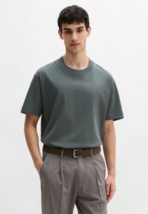Jonge man die een donker groen oversized T-shirt draagt, in een grijze geplooide broek met een bruine riem, staand met één hand in zijn zak.