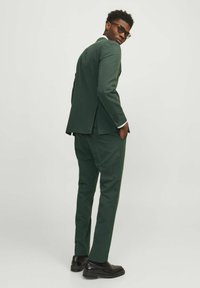 Traje ajustado de color verde oscuro con chaqueta de una sola abertura, puños de camisa blanca visibles y mocasines negros. El modelo posa con las manos en los bolsillos.