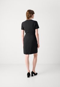 Black dress with short sleeves, fitted waist, and a zipper at the back. Smooth fabric and knee-length design. Worn with black, heeled shoes.
