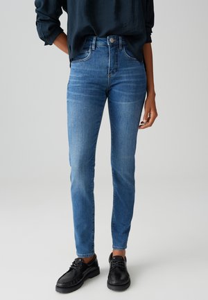 Osoba ubrana w niebieskie, dopasowane dżinsy o średnim odcieniu, czarne sznurowane buty oraz granatową koszulę z długim rękawem wpuszczoną w spodnie na jasnoszarym tle.