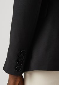 Rękaw czarnego blezera z czterema czarnymi guzikami i dopasowanym fasonem o gładkiej fakturze. Tkanina jest strukturalna i o czystym kroju.