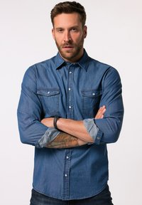 Camisa de mezclilla en azul oscuro con mangas largas, frente abotonado, dos bolsillos en el pecho y acentos de puño en un azul más claro. Ajuste entallado.