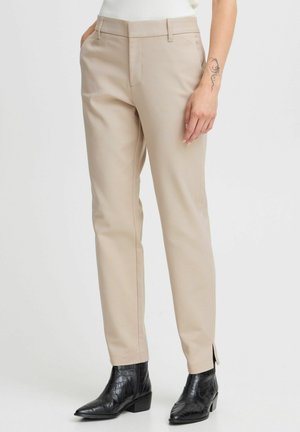 Pantalon beige ajusté avec poches latérales, coupe slim et fentes latérales discrètes à l'ourlet, associé à des bottines noires en cuir à motif crocodile.