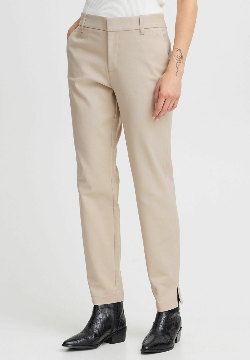 Pantalon beige ajusté avec poches latérales, coupe slim et fentes latérales discrètes à l'ourlet, associé à des bottines noires en cuir à motif crocodile.