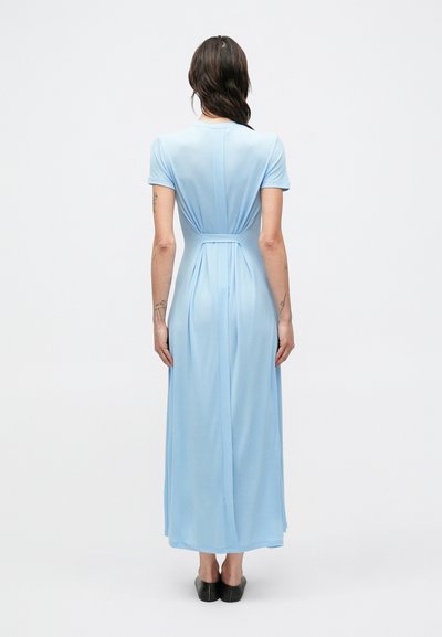 Svetlomodré maxi šaty s krátkym rukávom a plynulou siluetou, ktoré majú nariasený chrbát a hladkú textúru. Čierne topánky dopĺňajú tento vzhľad.