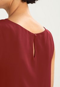 Mørkerød bluse med et glat, blødt stof og en ryg med nøglehulåbning, der er sikret med en matchende knap. Enkle linjer og minimal design.